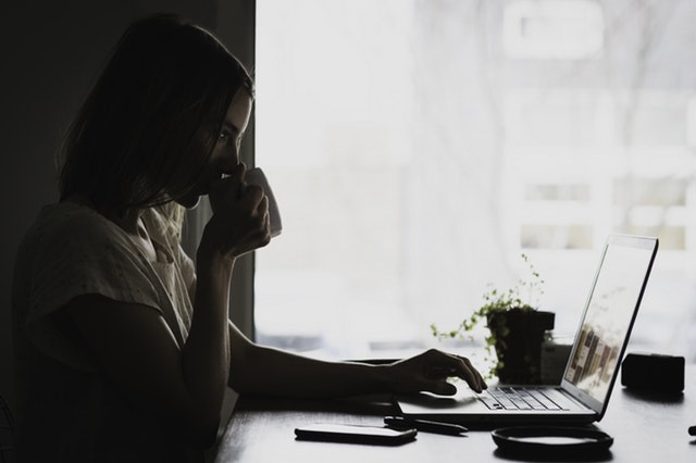 Woman sitting at a desk sipping a cofee and typing on her laptop