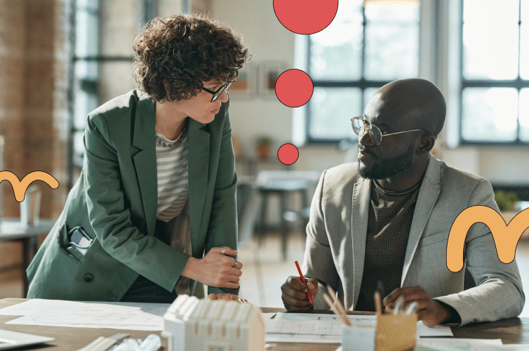 Two people at a desk. One sitting and one standing, looking at each other in conversation.